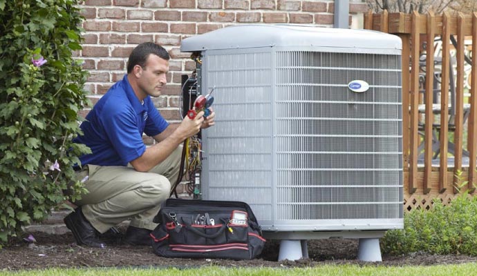 An HVAC technician in a blue uniform crouches down to repair the open panel of an outdoor Carrier air conditioning unit, with his tool bag on the ground.